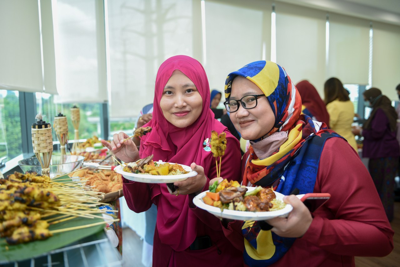 Two women enjoying plates of grilled skewers and festive dishes during Merdeka catering celebration at an office event.